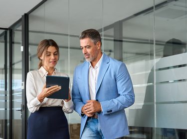 Executives team of two business people working talking using tablet tech walking in corporate office. Happy professional smiling businessman and businesswoman having conversation at work. Candid photo